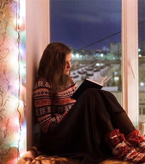 Woman reading in a window while wearing slippers and surrounded by a string of lights.