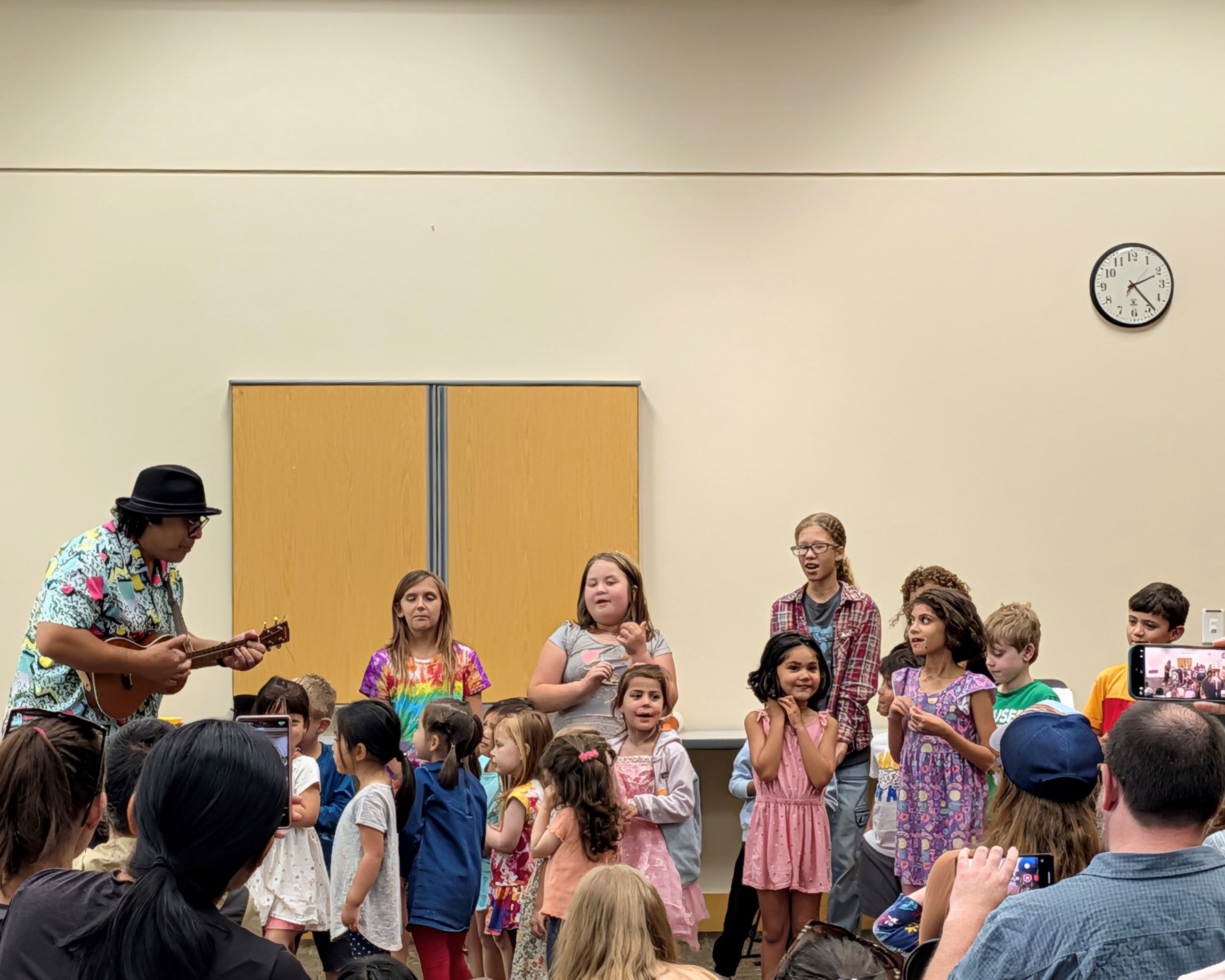 A musician playing a small guitar with a group of a dozen children, singing.